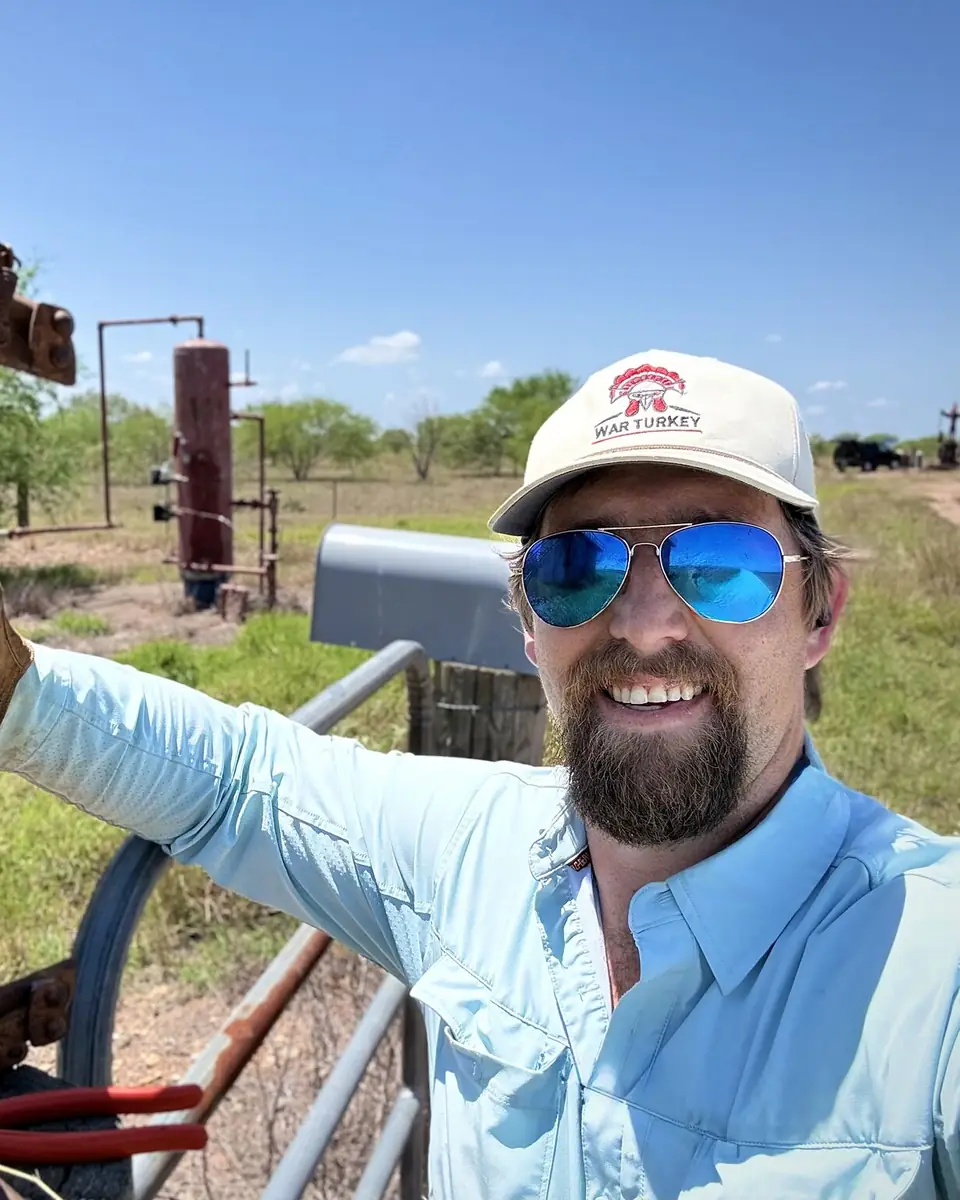 Reed Goodman in a War Turkey cap and sunglasses, holding a well jack by a South Texas wellhead.